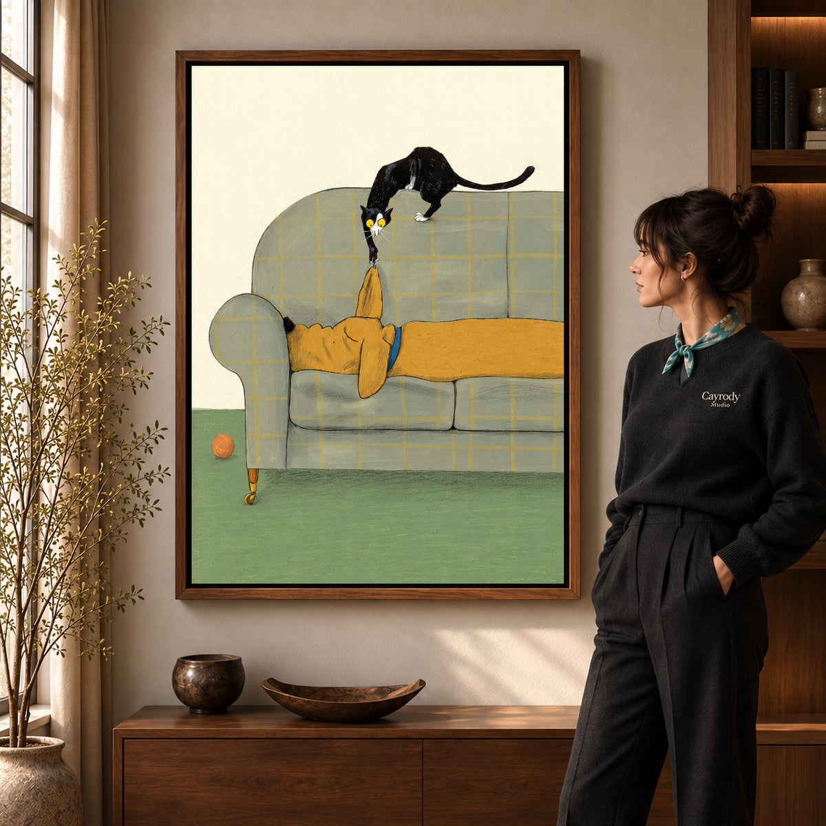Mischievous Sofa Kitten artwork in a walnut frame beside a standing woman in a warm room.