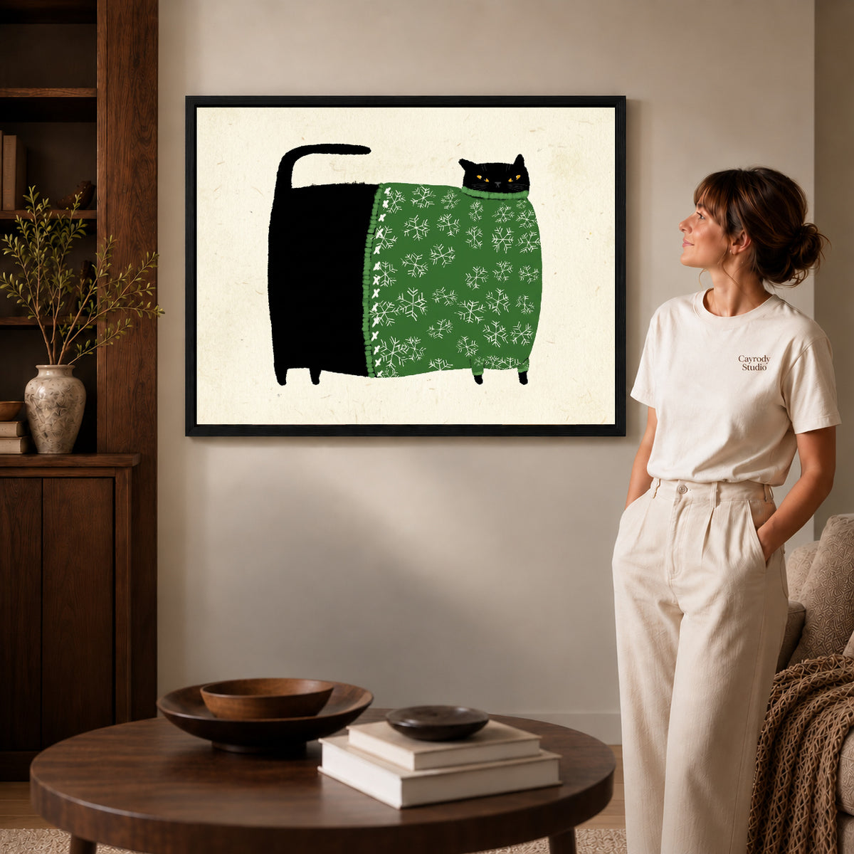 Black framed snowflake sweater cat artwork beside a woman in a warm neutral living room.