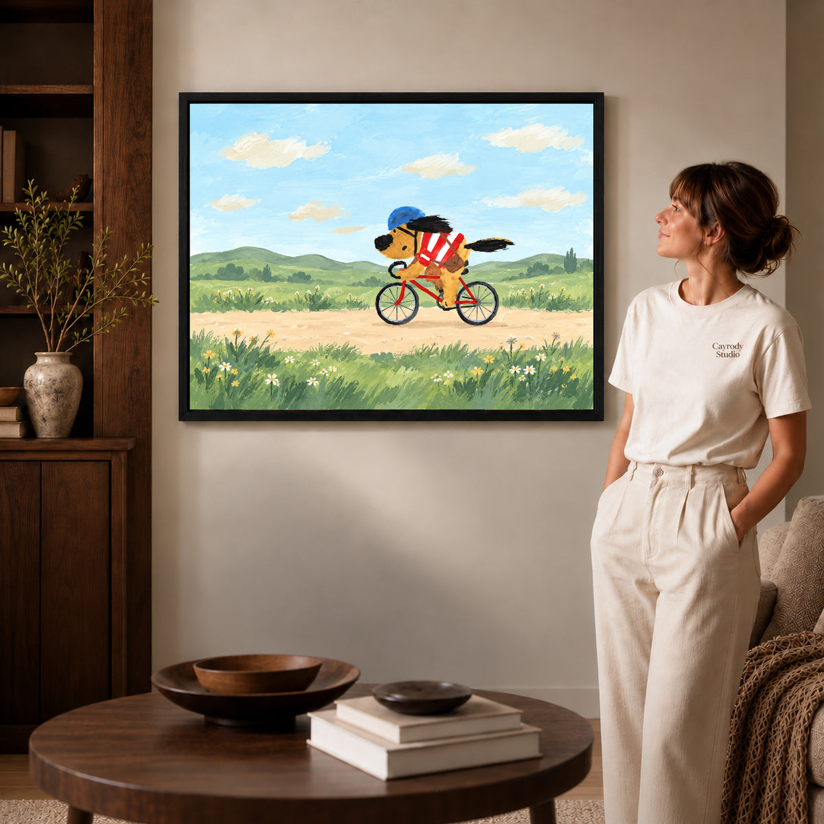 Black framed country cycling dog artwork beside a woman in a warm neutral living room.