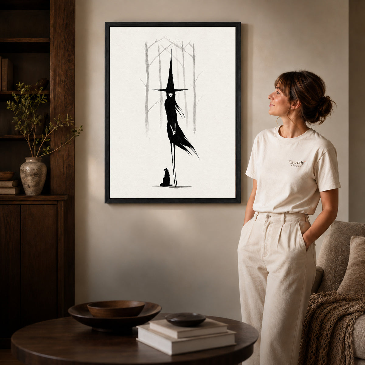 Black framed tall witch companion artwork beside a woman in a warm neutral living room.