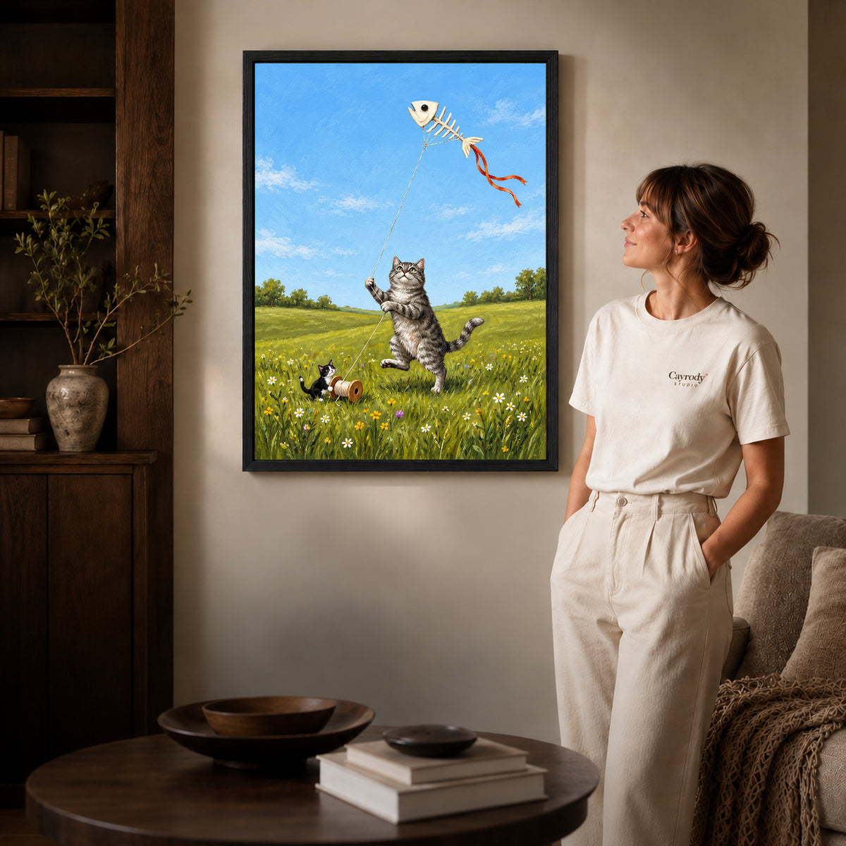 Black framed kitten kite friends artwork beside a woman in a warm neutral living room.