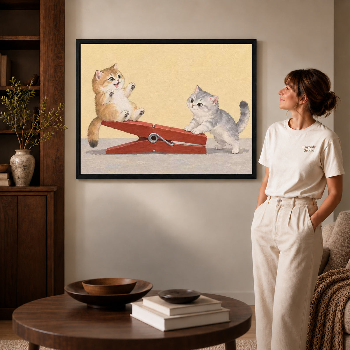 Black framed clothespin kitten friends artwork beside a woman in a warm neutral living room.
