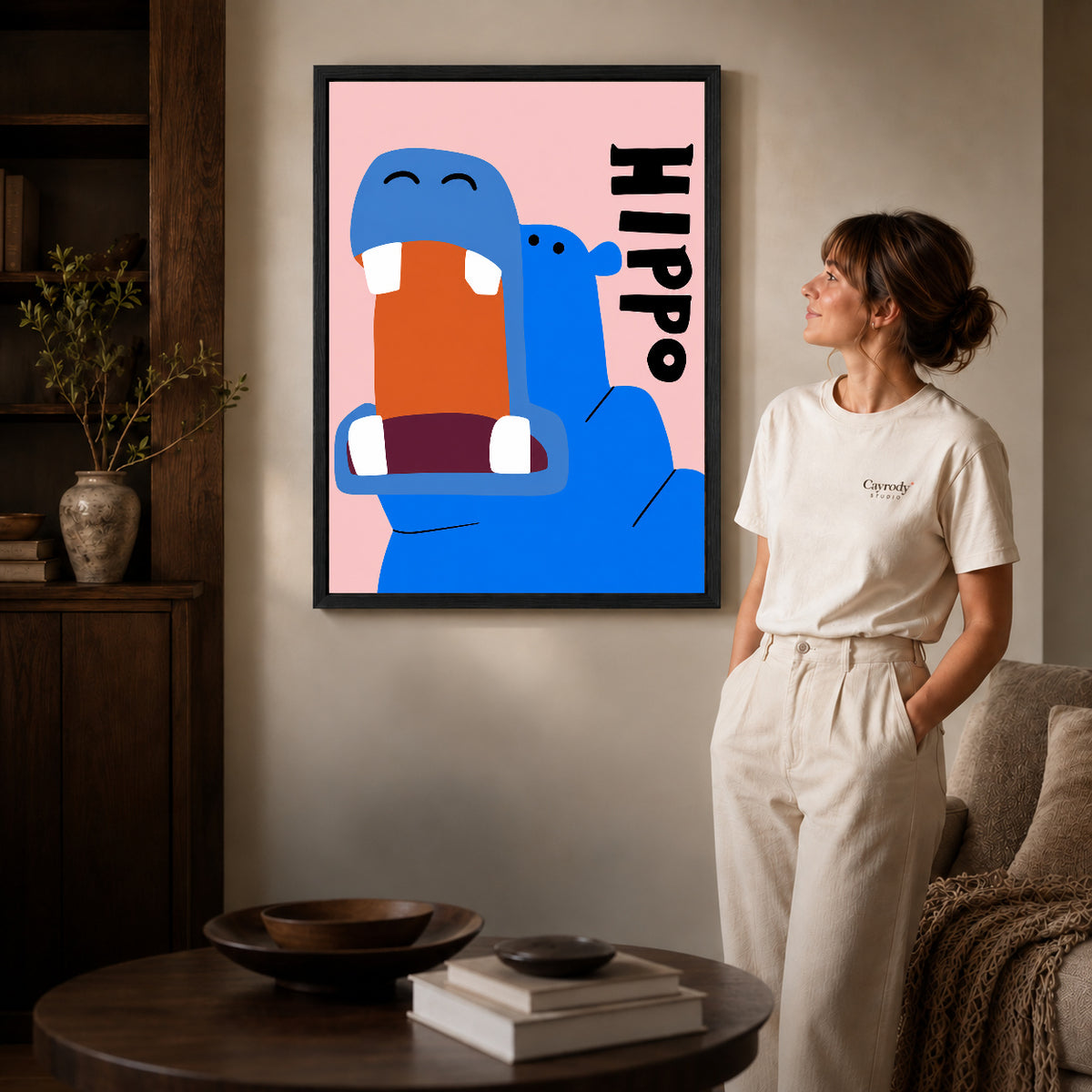 Black framed happy blue hippo artwork beside a woman in a warm neutral living room.