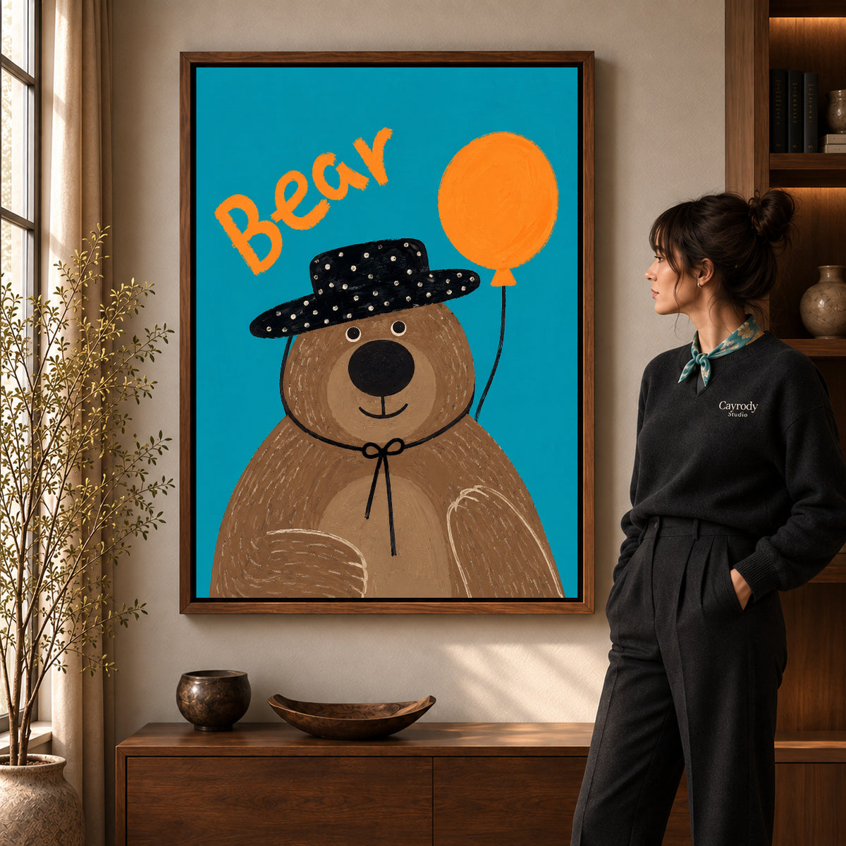 Balloon Bear Portrait artwork in a walnut frame beside a standing woman in a warm room.