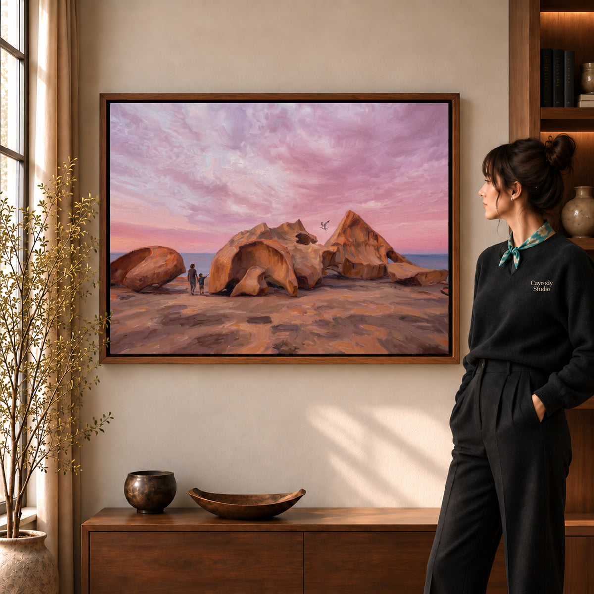 Coastal Rock Sunset artwork in a walnut frame beside a standing woman in a warm room.