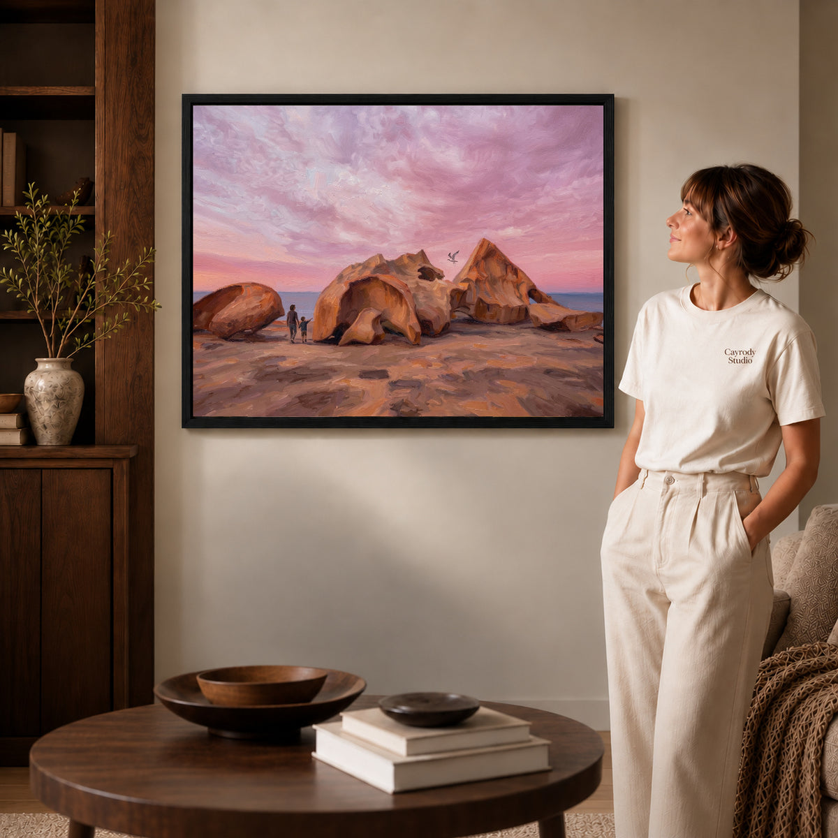 Black framed coastal rock sunset artwork beside a woman in a warm neutral living room.