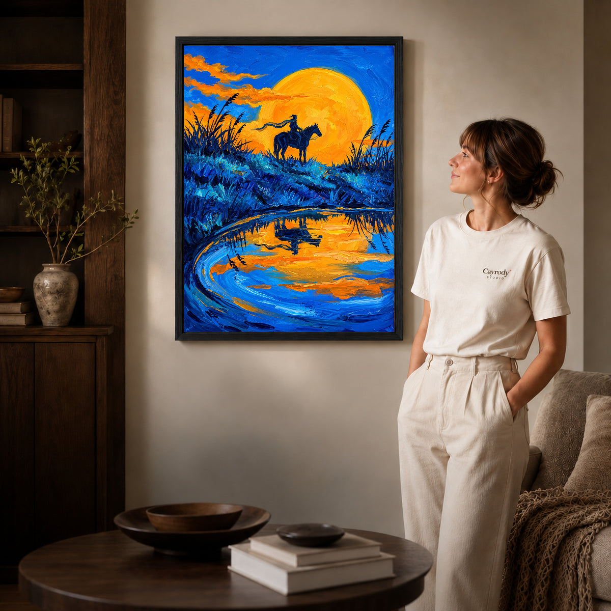 Black framed golden moon rider artwork beside a woman in a warm neutral living room.