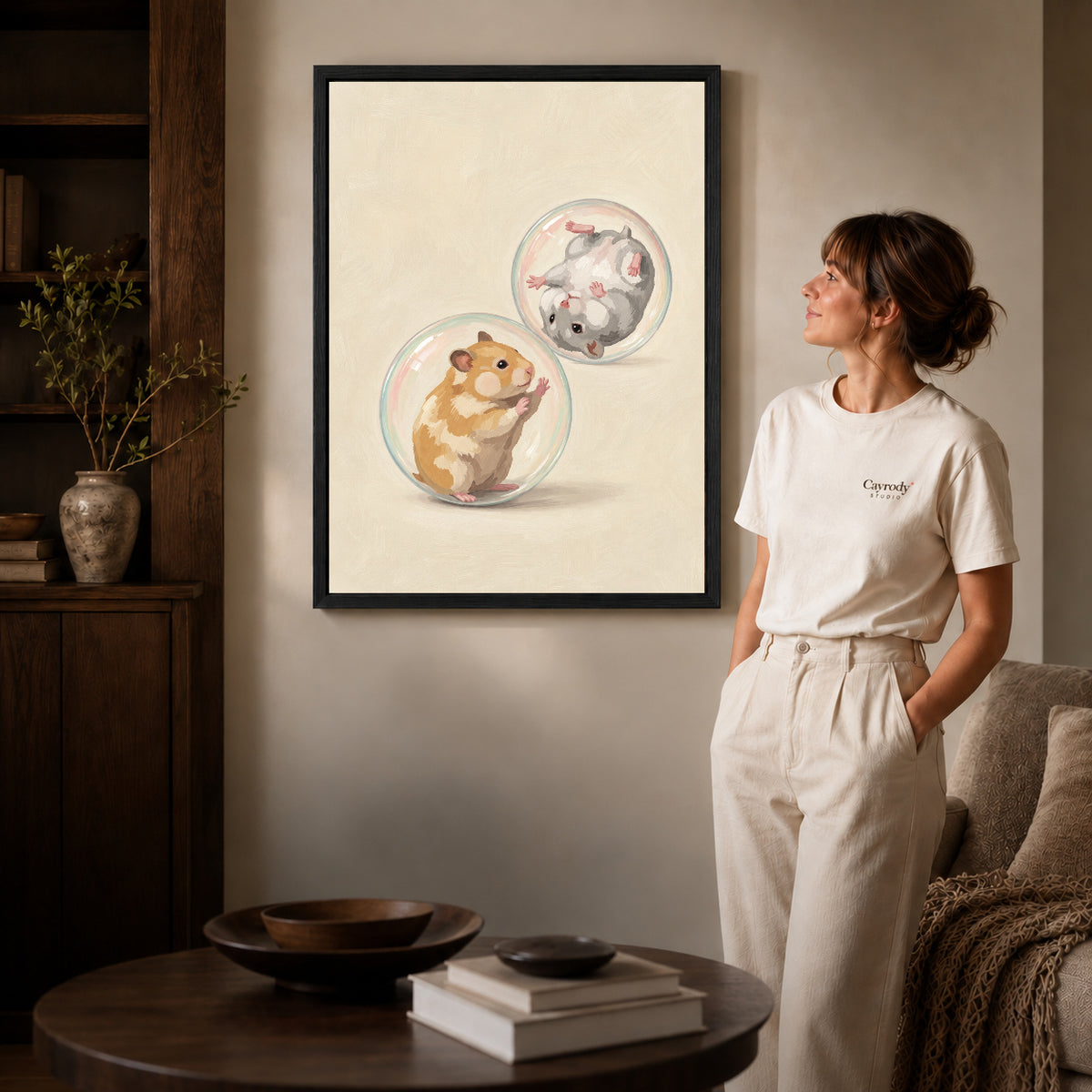 Black framed tumbling hamster friends artwork beside a woman in a warm neutral living room.