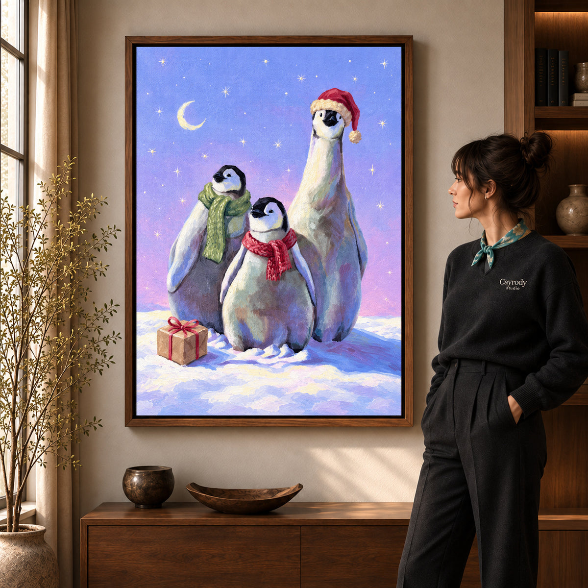 Christmas Penguin Trio artwork in a walnut frame beside a standing woman in a warm room.