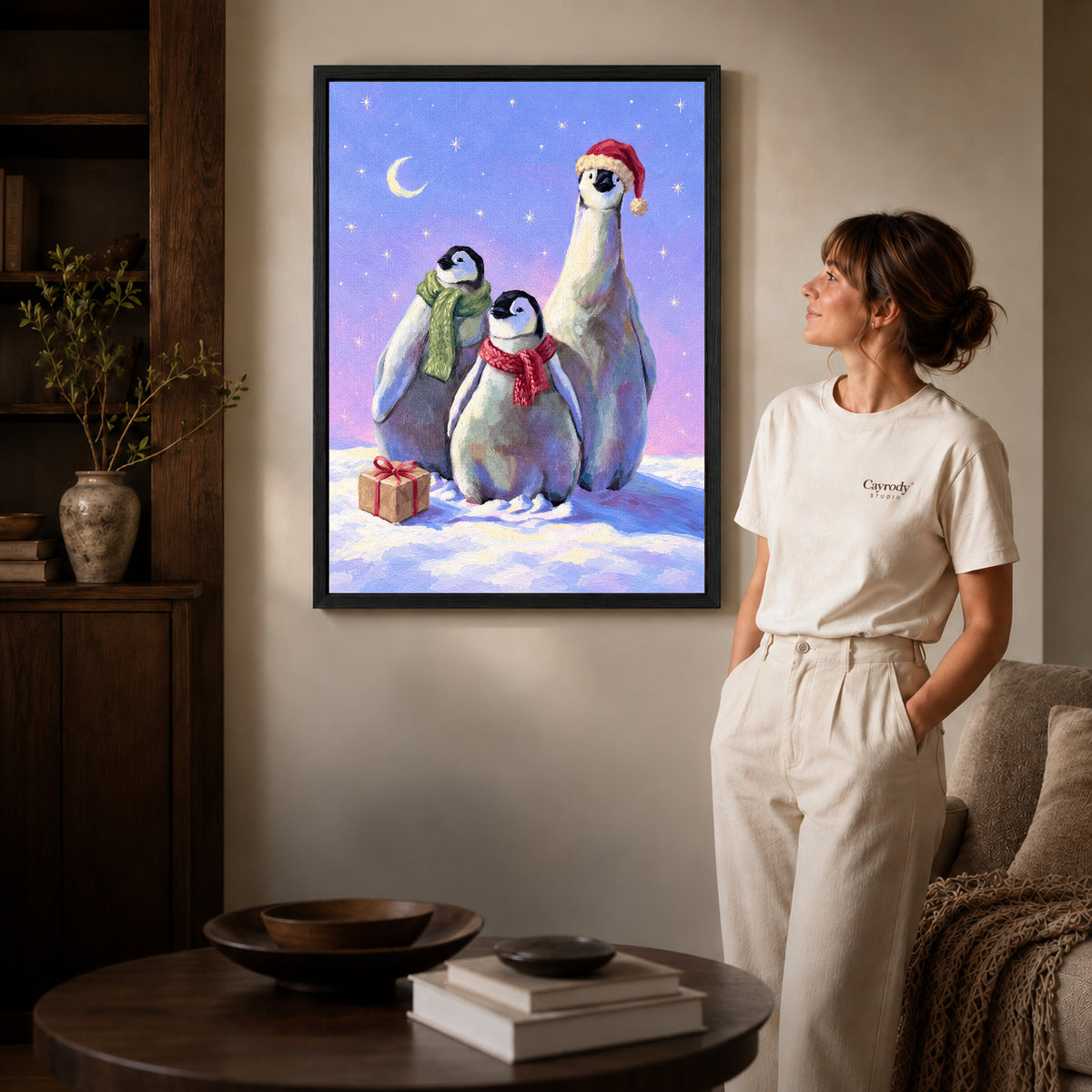 Black framed christmas penguin trio artwork beside a woman in a warm neutral living room.