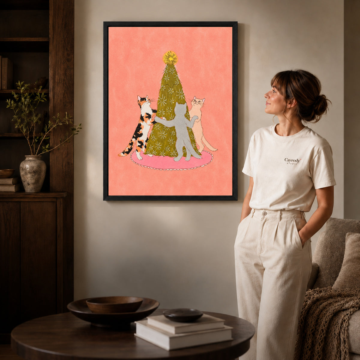 Black framed christmas tree cat dance artwork beside a woman in a warm neutral living room.