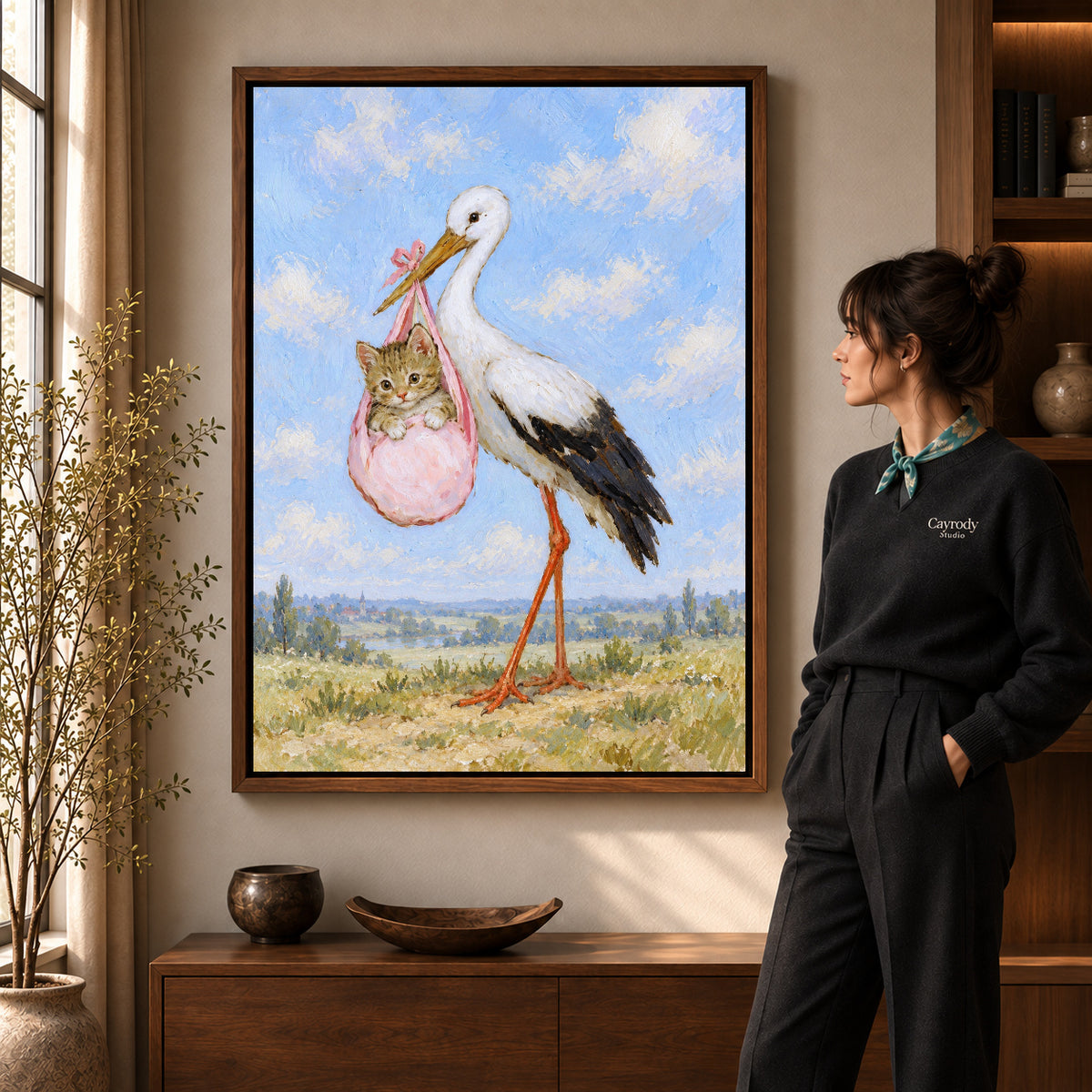 Stork Kitten Delivery artwork in a walnut frame beside a standing woman in a warm room.