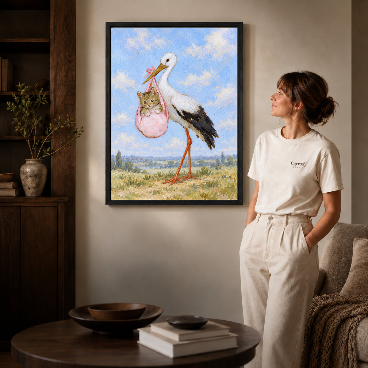 Black framed stork kitten delivery artwork beside a woman in a warm neutral living room.