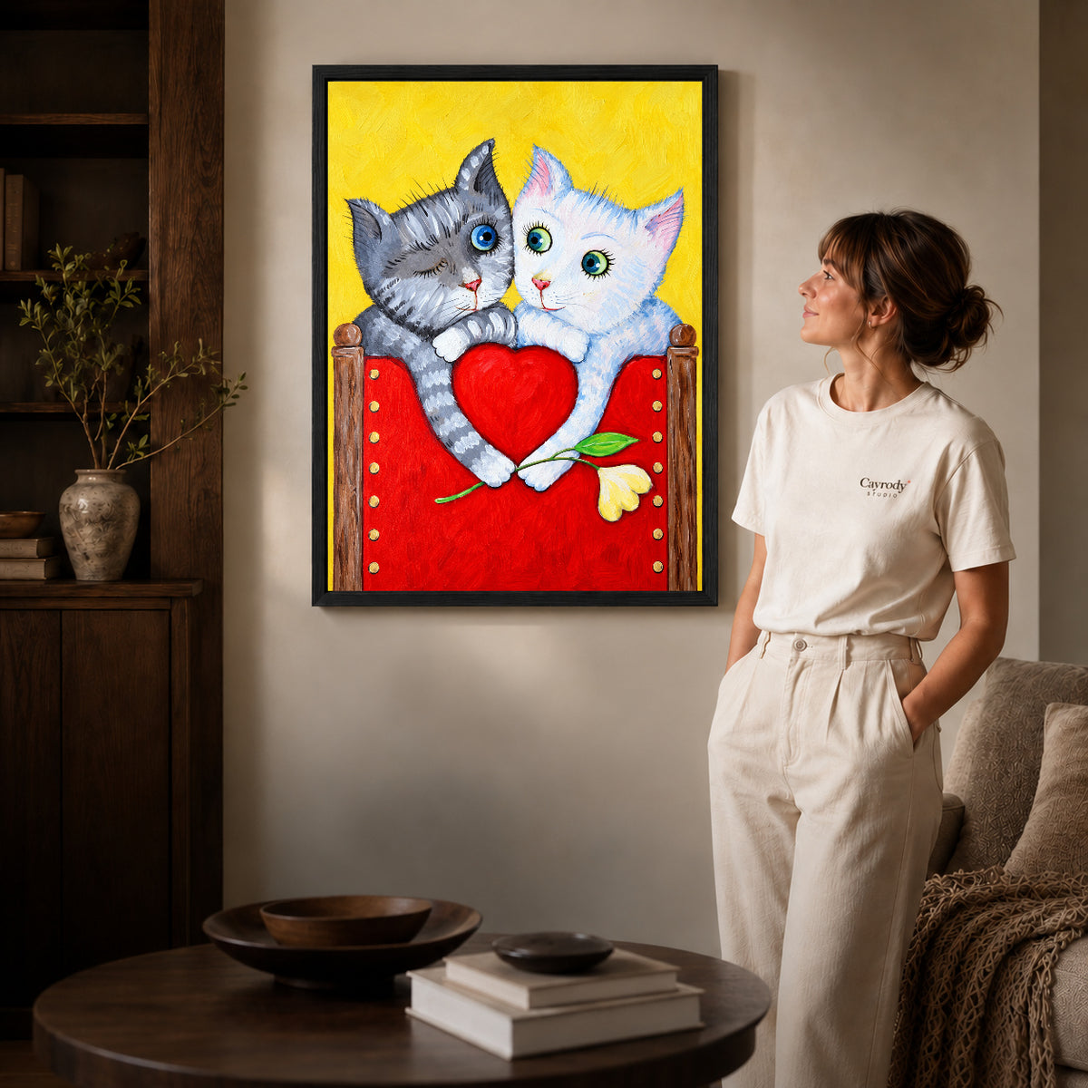 Black framed heart holding kittens artwork beside a woman in a warm neutral living room.