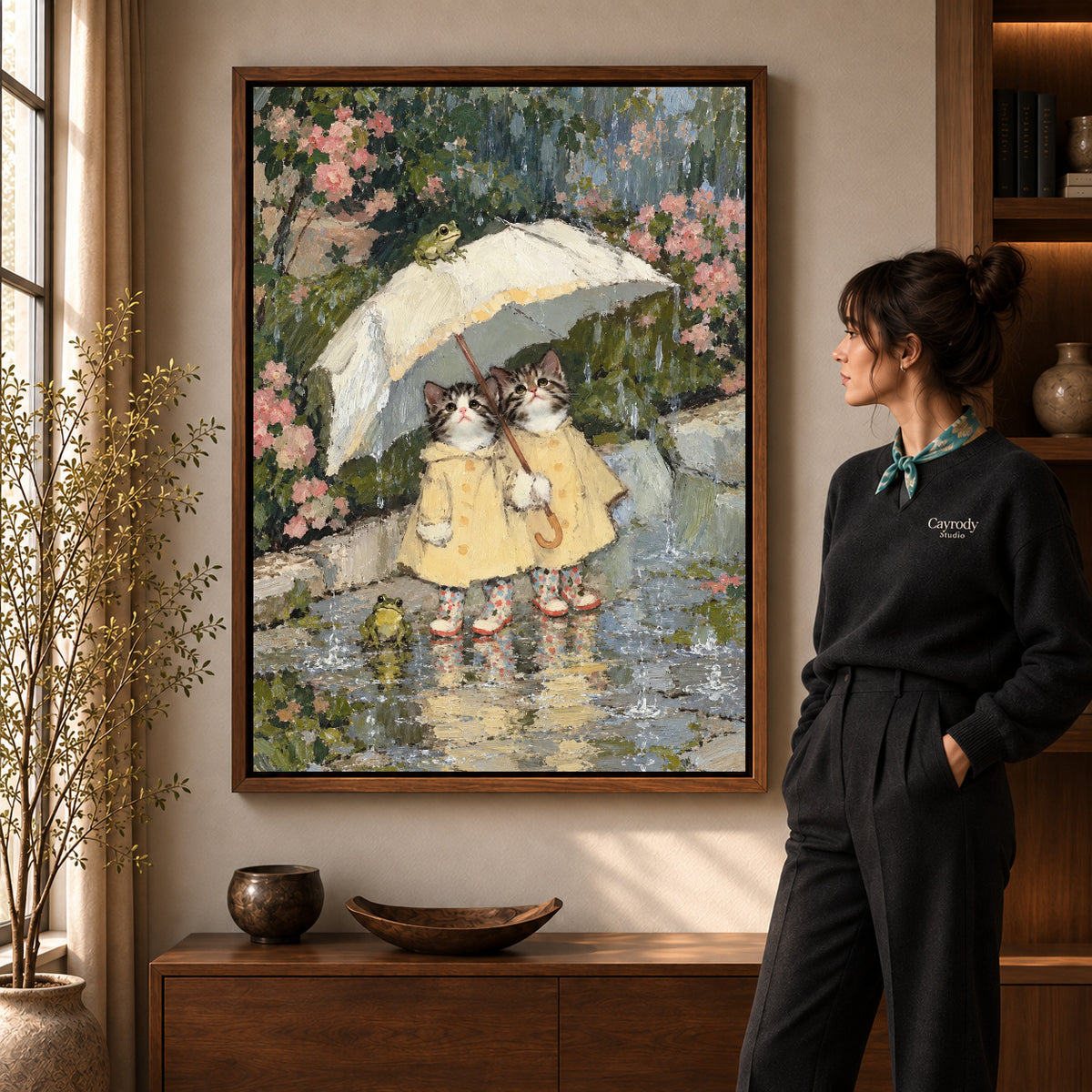 Raincoat Kitten Friends artwork in a walnut frame beside a standing woman in a warm room.