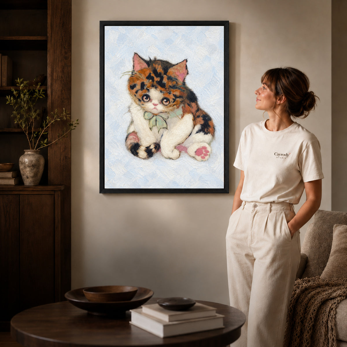 Black framed calico bow kitten artwork beside a woman in a warm neutral living room.