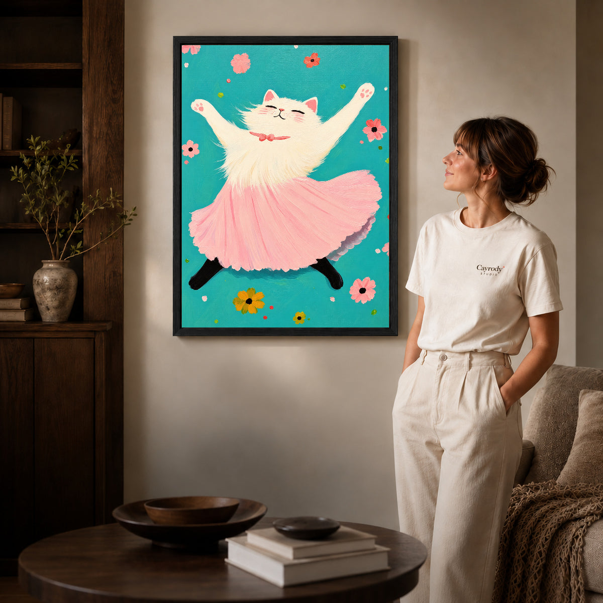 Black framed joyful ballet cat artwork beside a woman in a warm neutral living room.