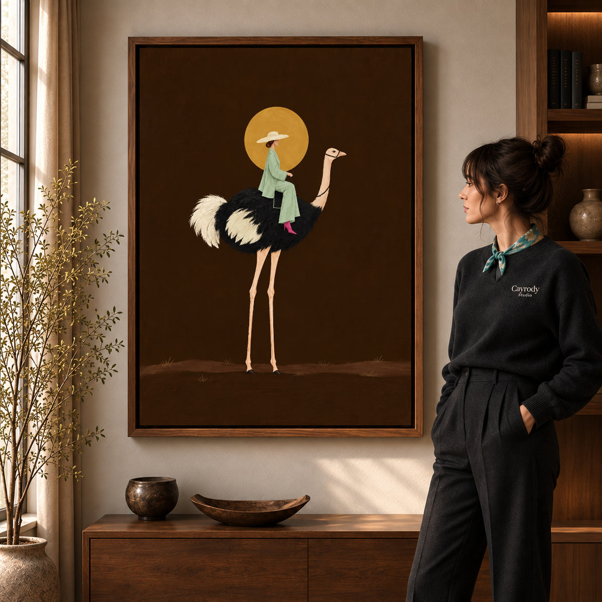 Moonlit Ostrich Rider artwork in a walnut frame beside a standing woman in a warm room.