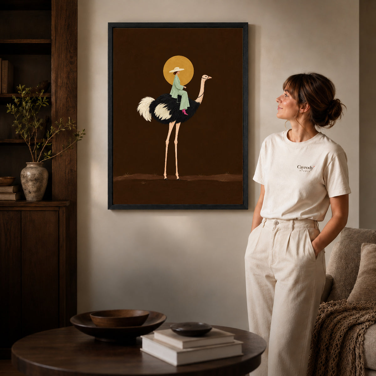 Black framed moonlit ostrich rider artwork beside a woman in a warm neutral living room.