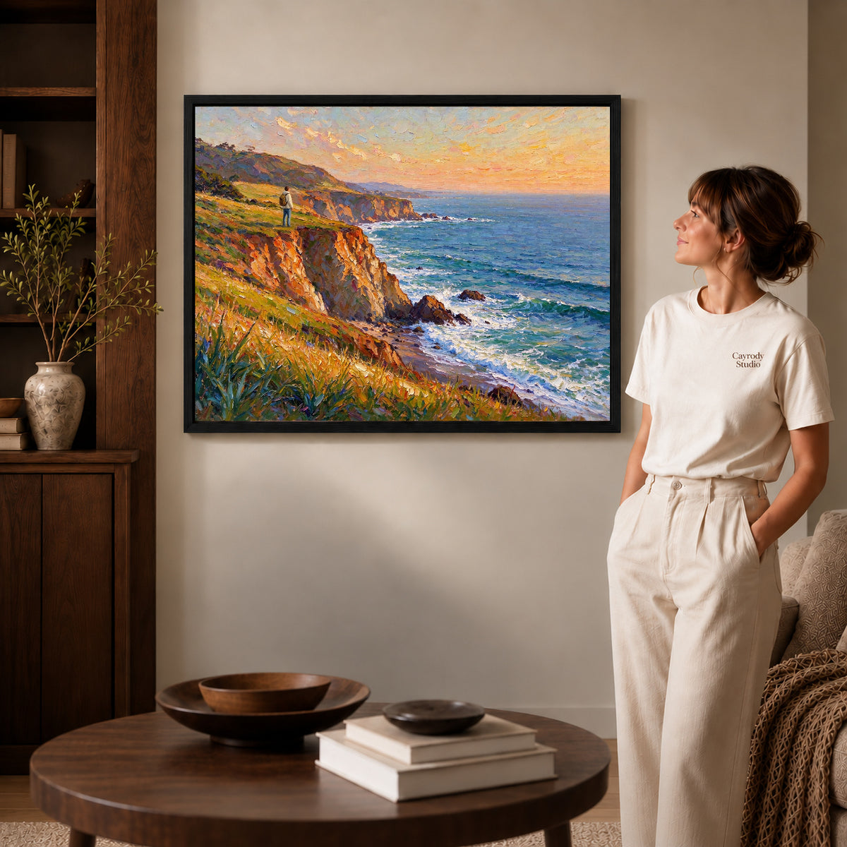 Black framed sunset coastal wanderer artwork beside a woman in a warm neutral living room.