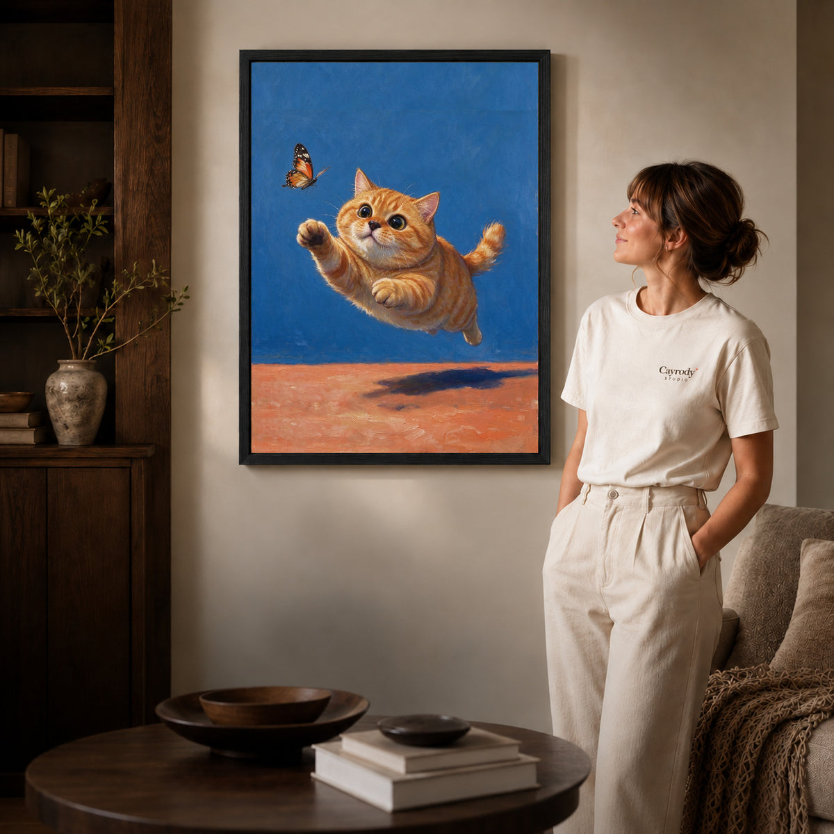 Black framed butterfly chasing kitten artwork beside a woman in a warm neutral living room.
