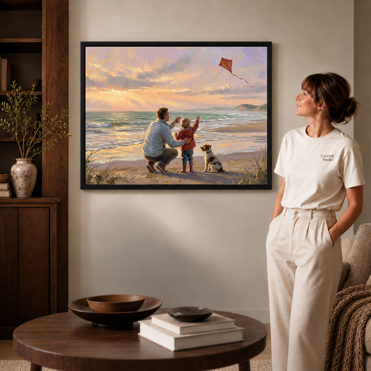 Black framed family beach kite artwork beside a woman in a warm neutral living room.