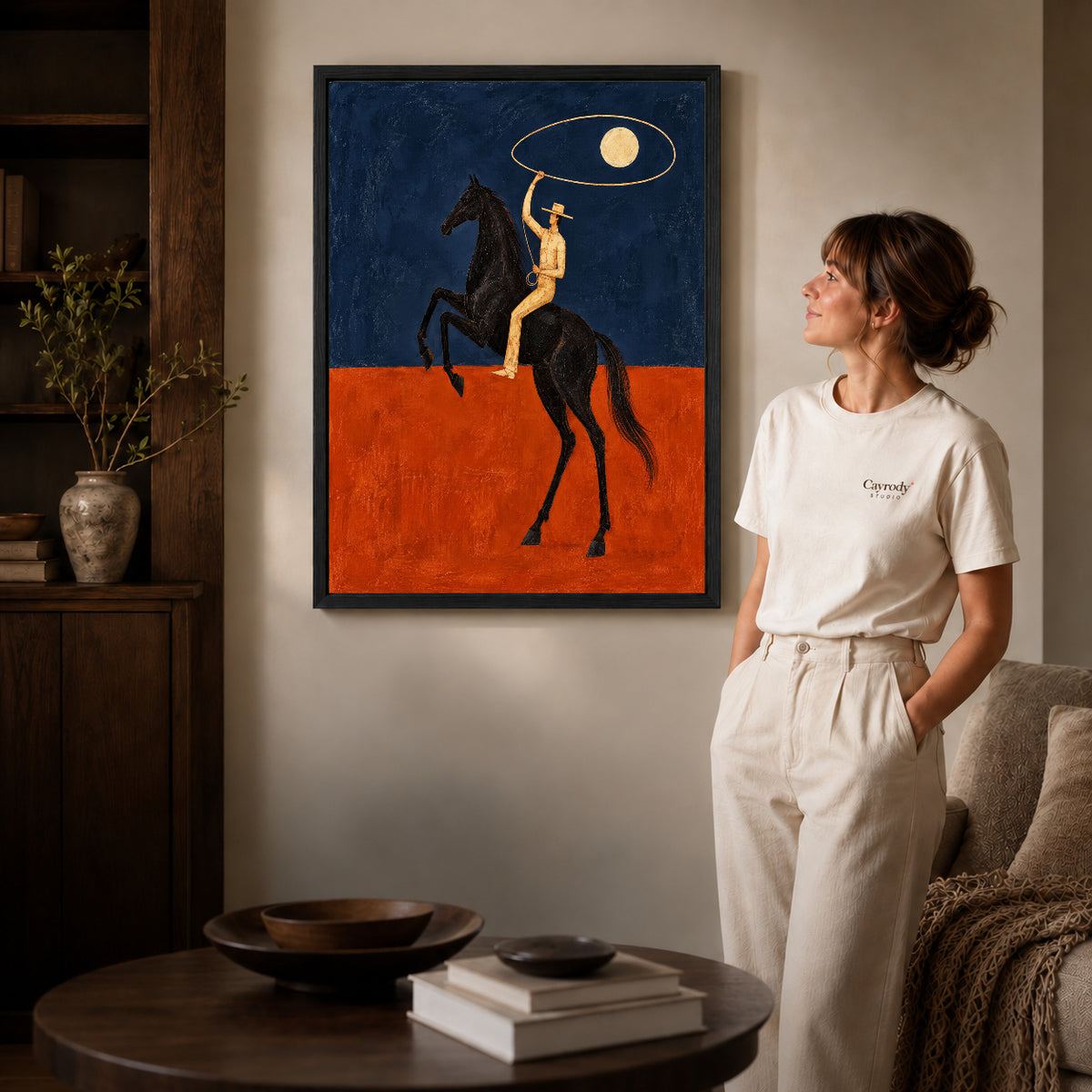 Black framed moon lasso rider artwork beside a woman in a warm neutral living room.