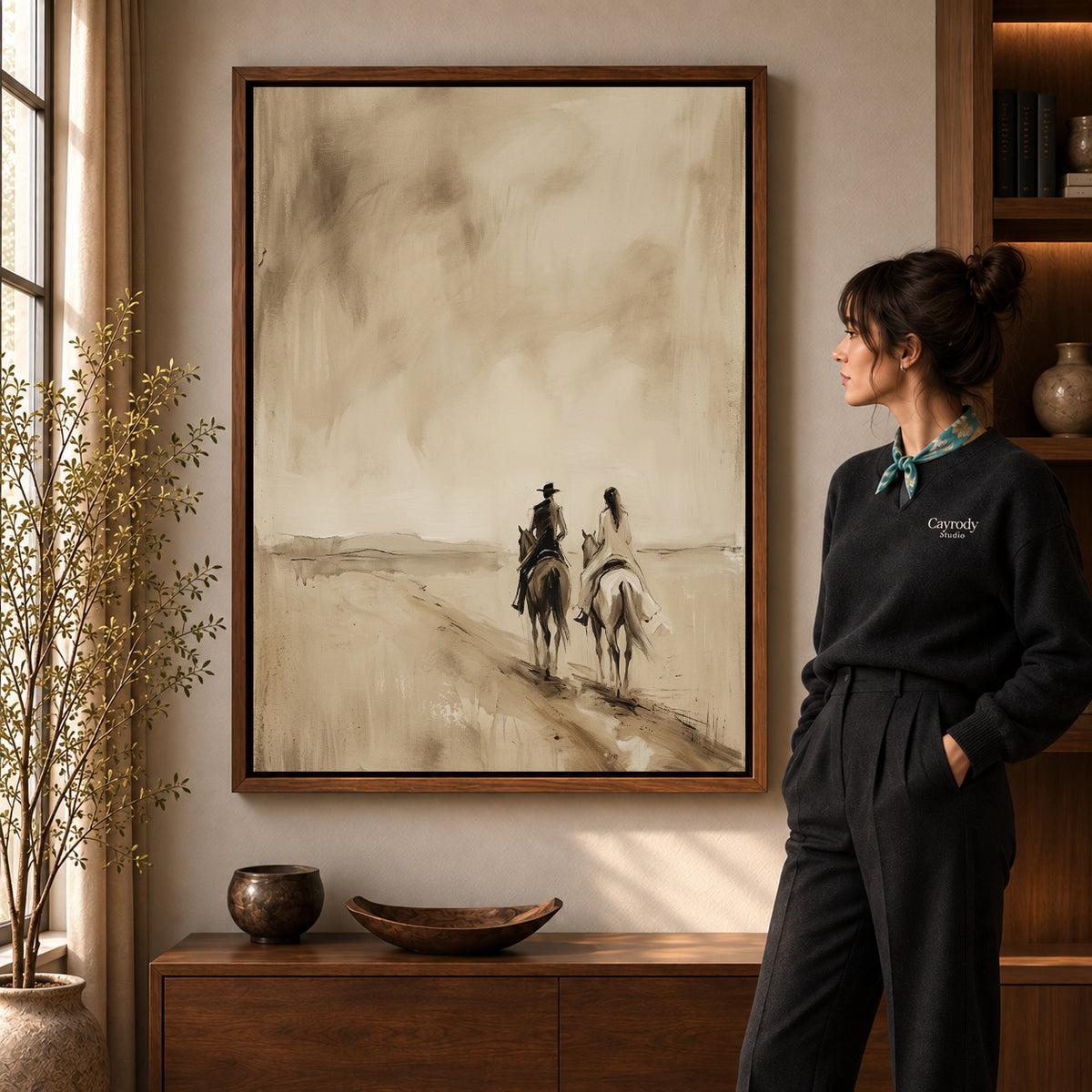Quiet Western Riders artwork in a walnut frame beside a standing woman in a warm room.