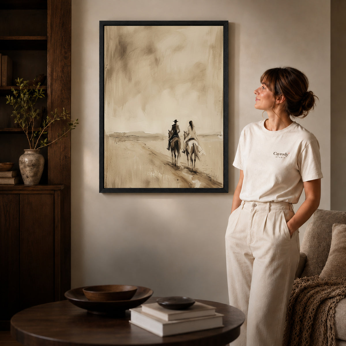 Black framed quiet western riders artwork beside a woman in a warm neutral living room.