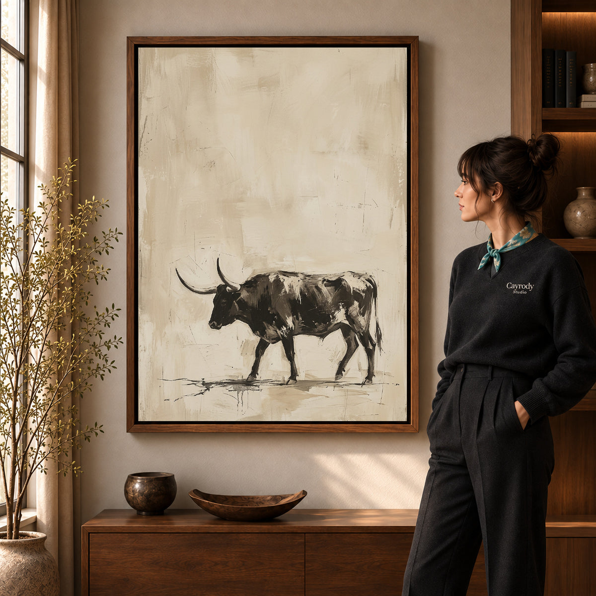 Neutral Longhorn Bull artwork in a walnut frame beside a standing woman in a warm room.