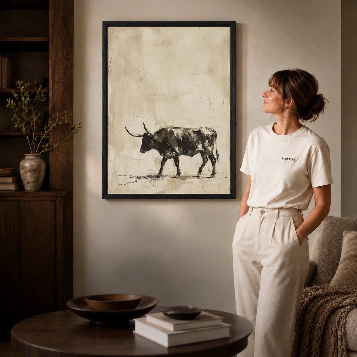 Black framed neutral longhorn bull artwork beside a woman in a warm neutral living room.