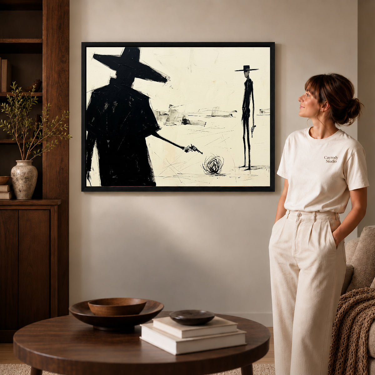 Black framed western desert standoff artwork beside a woman in a warm neutral living room.