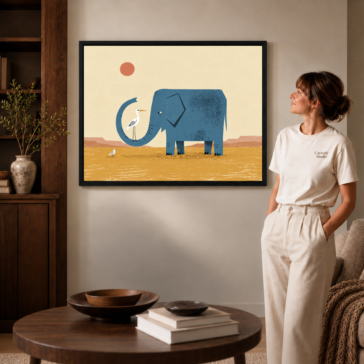 Black framed elephant bird friends artwork beside a woman in a warm neutral living room.