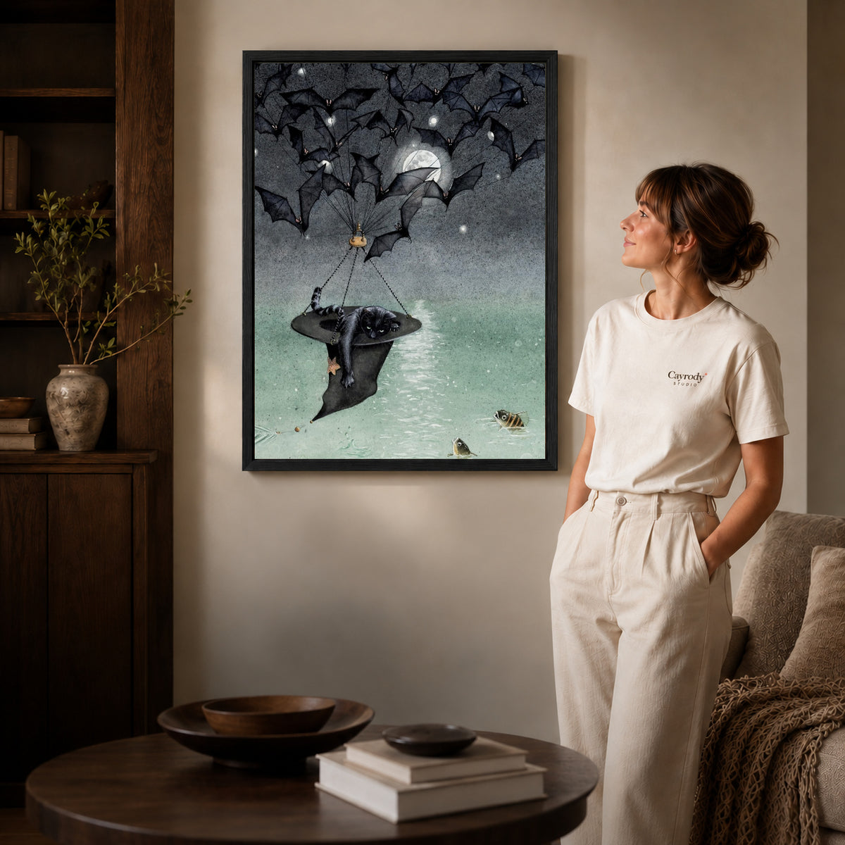 Black framed moonlit bat voyage artwork beside a woman in a warm neutral living room.