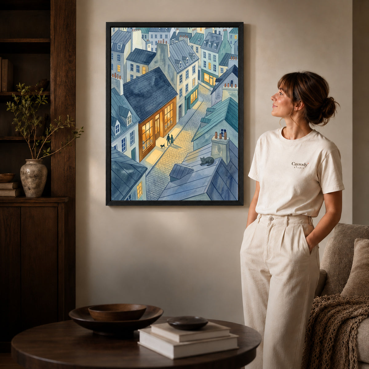 Black framed blue rooftop evening artwork beside a woman in a warm neutral living room.