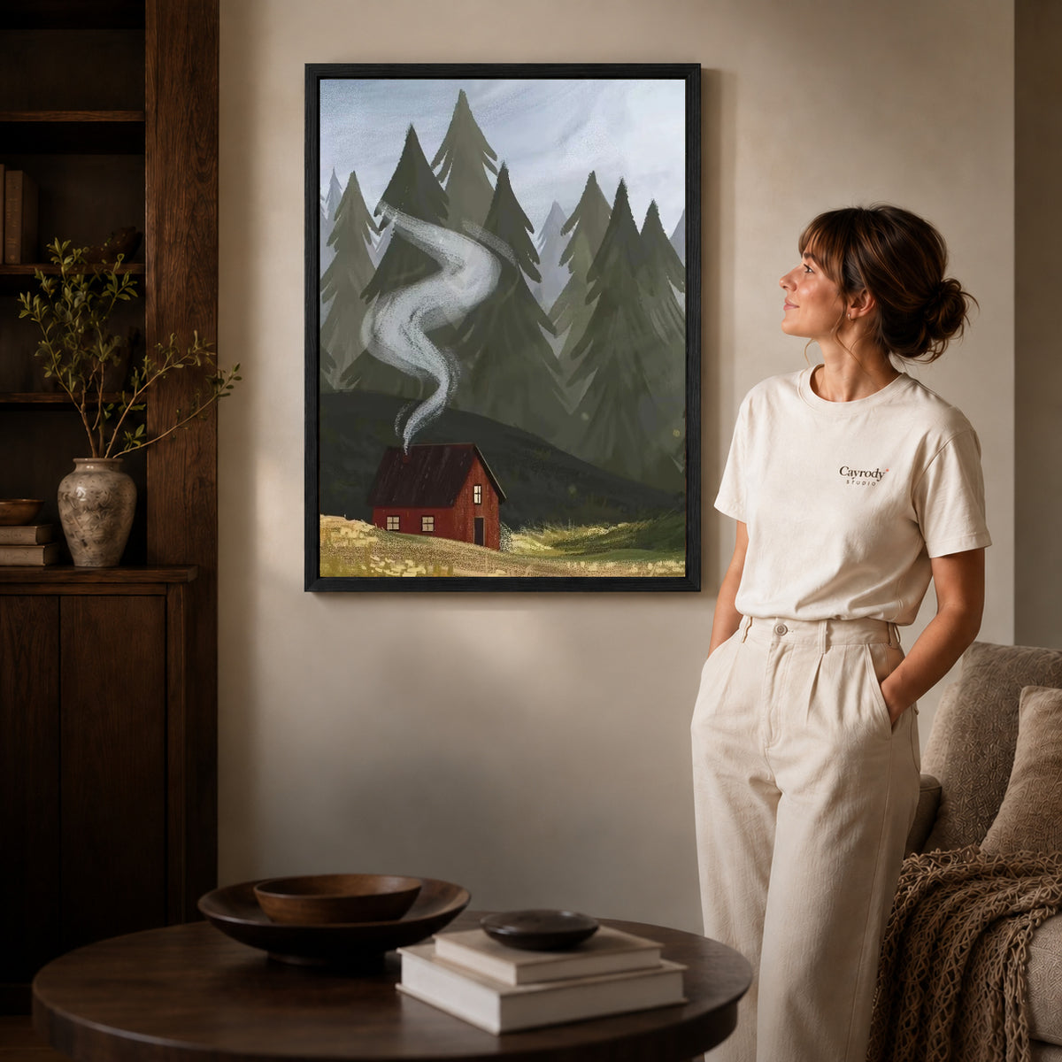 Black framed forest cabin smoke artwork beside a woman in a warm neutral living room.