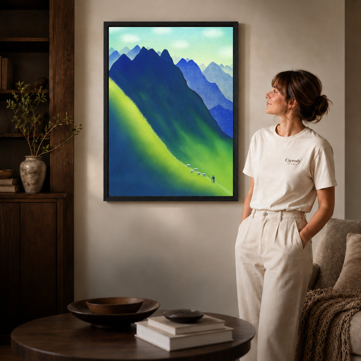 Black framed blue mountain canvas art displayed beside a woman in a warm neutral living room.