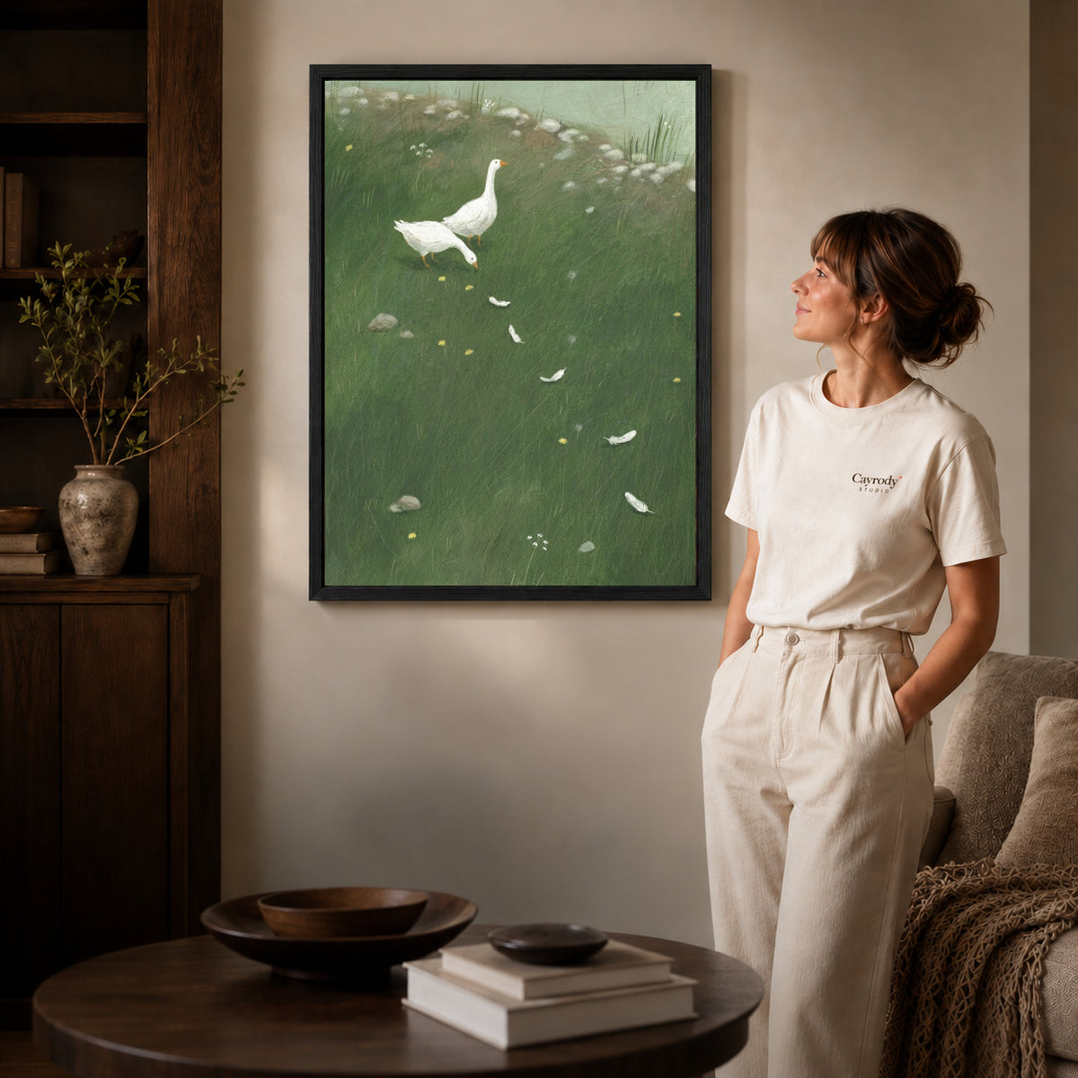 Black framed white goose meadow canvas art displayed beside a woman in a warm neutral living room.