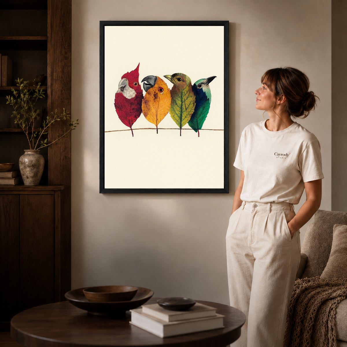 Black framed autumn leaf birds artwork beside a woman in a warm neutral living room.