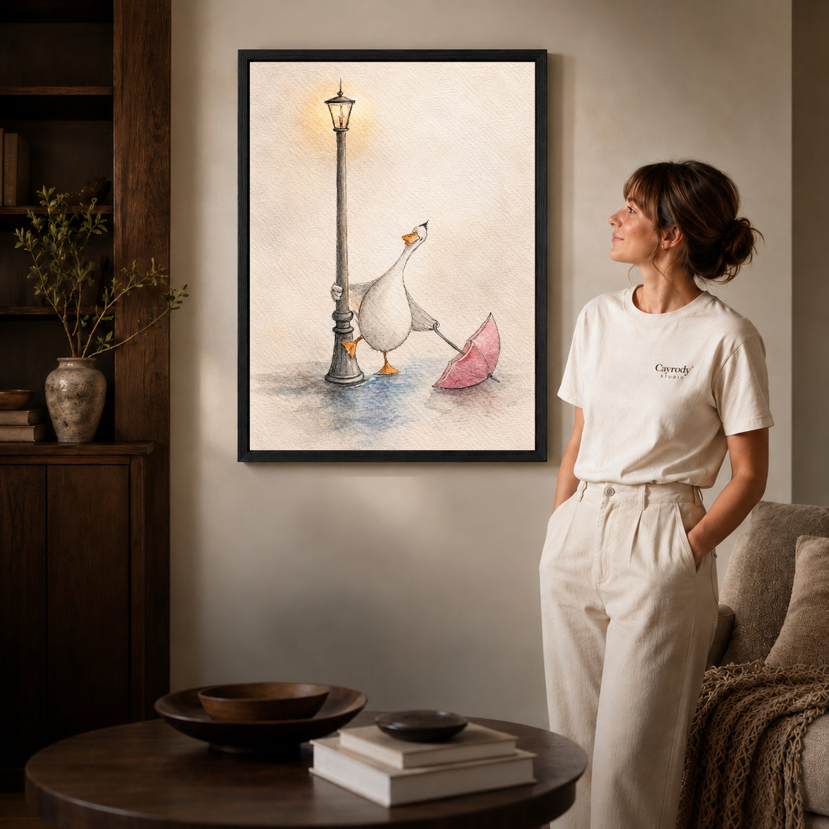 Black framed rainy lamppost goose artwork beside a woman in a warm neutral living room.